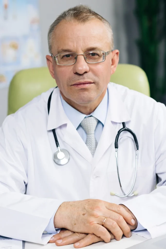 Male physician in a white coat with a stethoscope seated in a medical office.